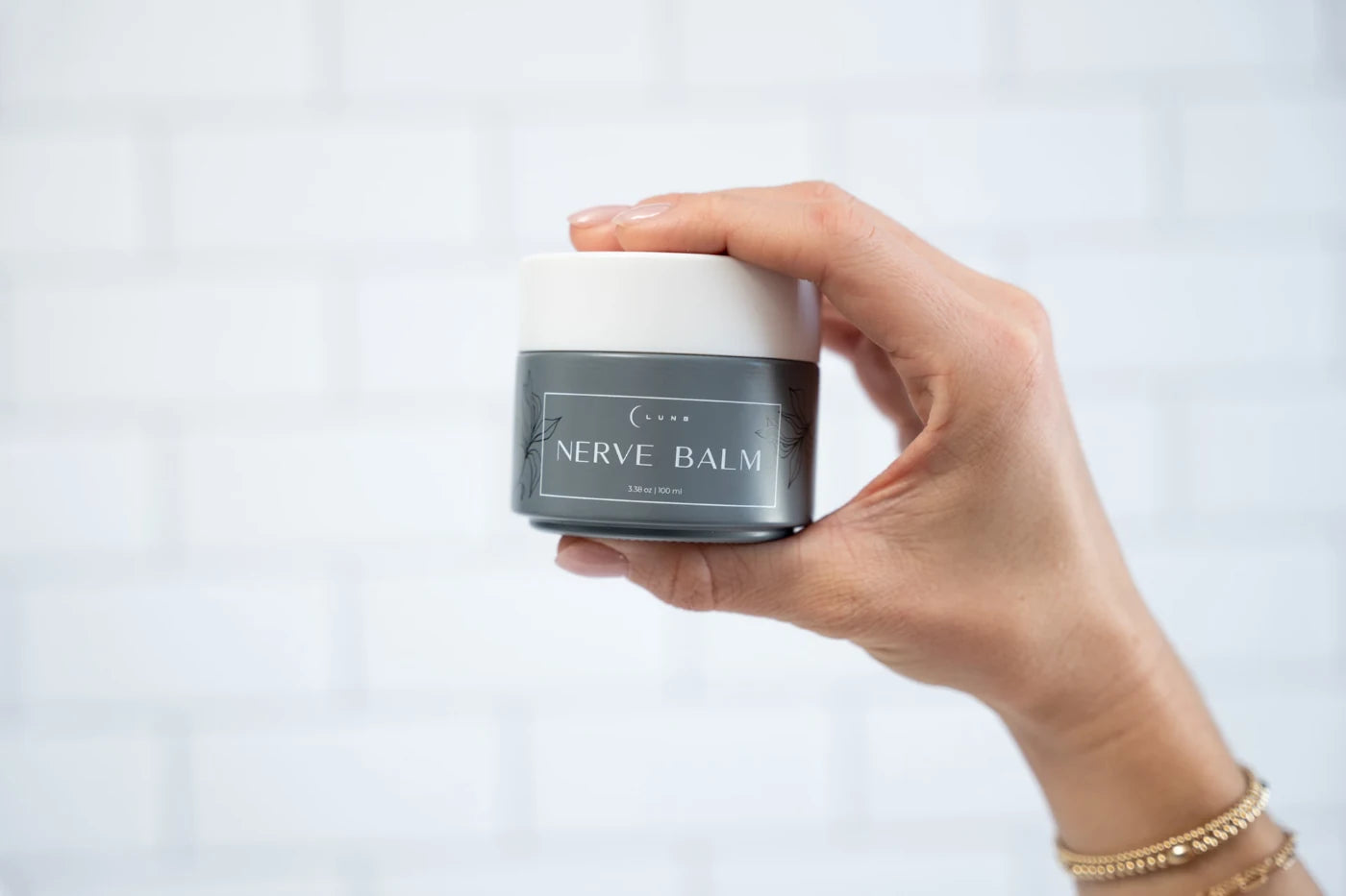 A hand holding a jar of Lune Nerve Balm against a white tile wall.