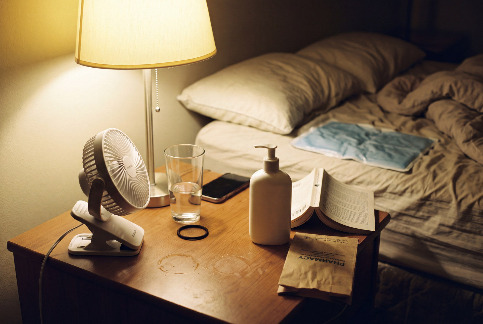 A bedside table at night with a small clip fan running, a glass of water, a phone face down, a plain bottle of lotion and a cooling gel pad on the sheet behind it.